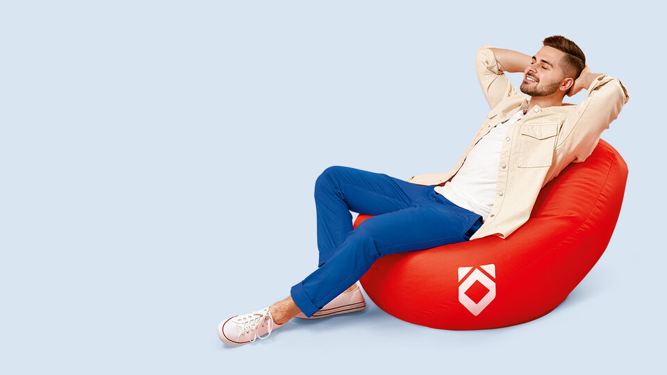 Junger Mann sitzt entspannt in einem roten Sitzsack mit gefalteten Armen und lächelt, vor einem hellblauen Hintergrund.