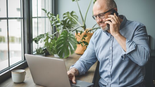 Mann mit Brille spricht am Telefon, während er an einem Laptop arbeitet, umgeben von Pflanzen am Fenster.