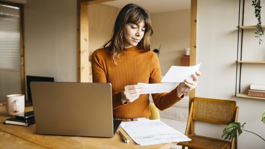 Frau mit langen Haaren in einem orangefarbenen Pullover schaut auf Dokumente am Schreibtisch, neben einem Laptop und einer Tasse.