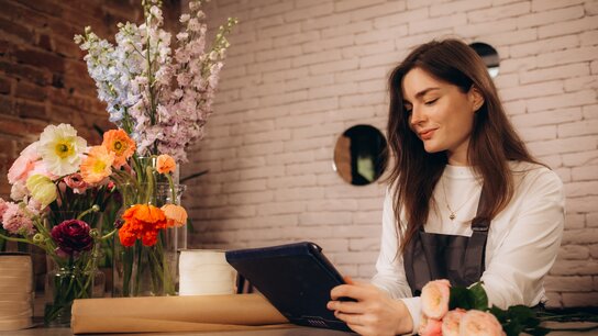 Floristin in einem Blumenladen, die auf ein Tablet schaut, umgeben von bunten Blumen und gedeckter Theke.