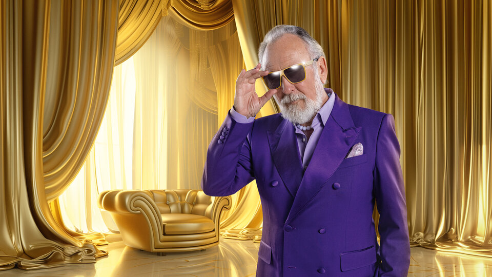 Mann mit grauem Bart und Sonnenbrille in lila Anzug, vor goldenen Vorhängen und einem goldenen Sofa.