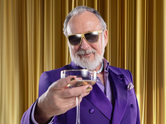 Ein älterer Mann mit Bart und Sonnenbrille in lila Anzug hält ein Glas Sekt vor goldenen Vorhängen.